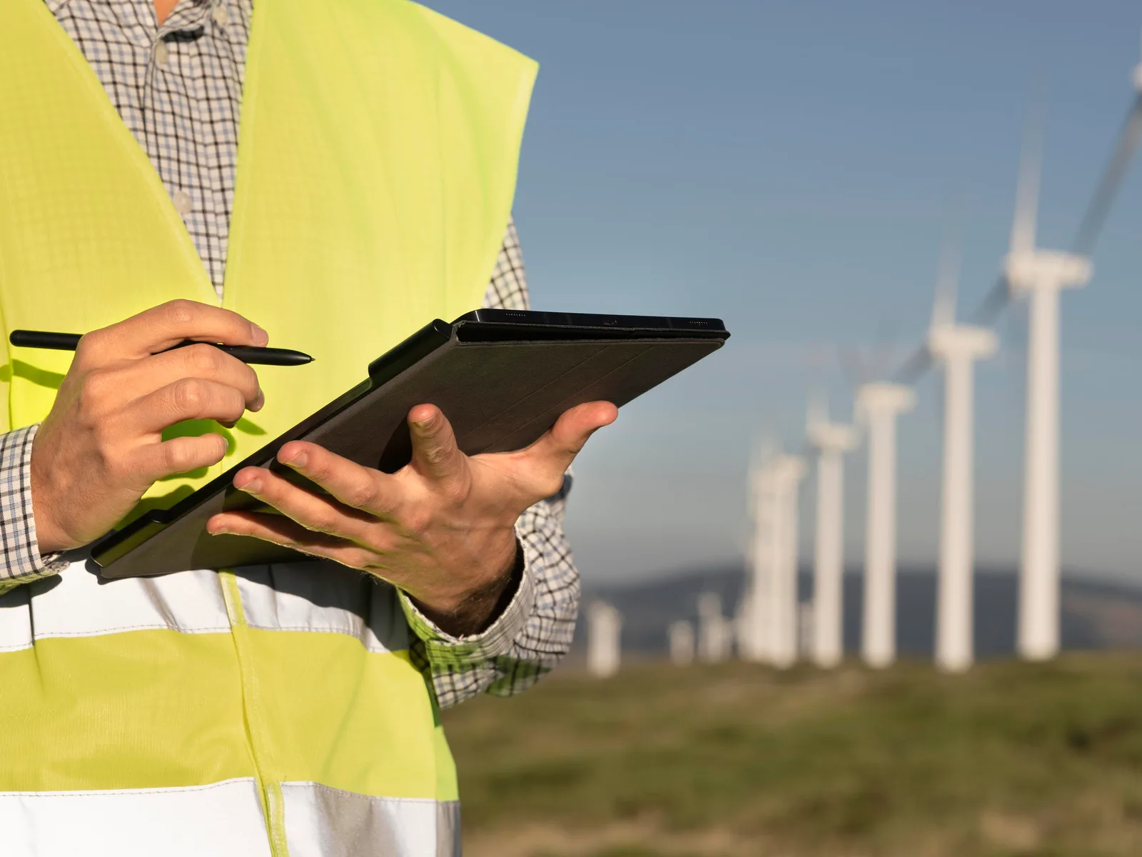 Técnico com colete de alta visibilidade faz anotações em um tablet diante de uma fileira de aerogeradores
