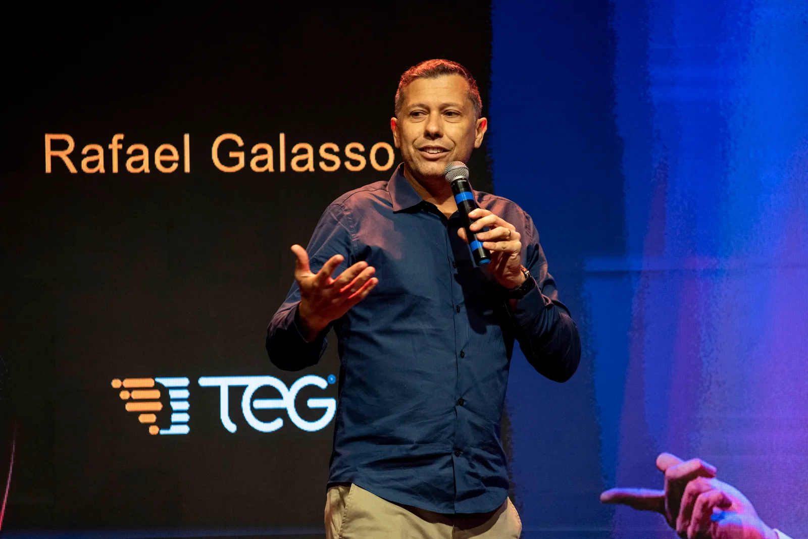 Rafael Galasso ao microfone em um evento da TEG, com seu nome e o logo TEG no telão ao fundo